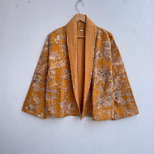 Puede incluir: Chaqueta estilo kimono de color amarillo mostaza con un estampado floral en beige y marrón. La chaqueta tiene un cuello chal y está colgada de una percha de madera. La chaqueta está abierta por delante, revelando un forro amarillo más claro.