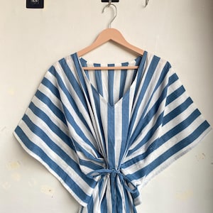 May include: A blue and white striped caftan dress with a V-neck and tie waist, hanging on a wooden hanger. The dress features wide, vertical stripes and a loose, flowing design, perfect for warm weather.