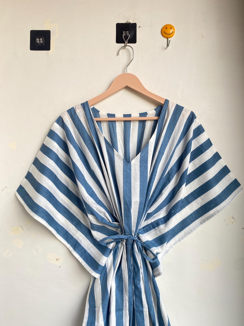 May include: A blue and white striped caftan dress with a V-neck and tie waist, hanging on a wooden hanger. The dress features wide, vertical stripes and a loose, flowing design, perfect for warm weather.