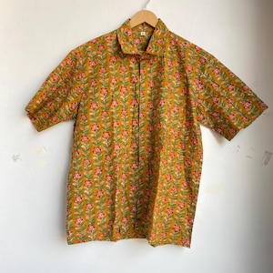 May include: A short-sleeved button-down shirt in a mustard yellow color, adorned with a repeating floral pattern in pink and green. The shirt is hanging on a wooden hanger against a white wall.