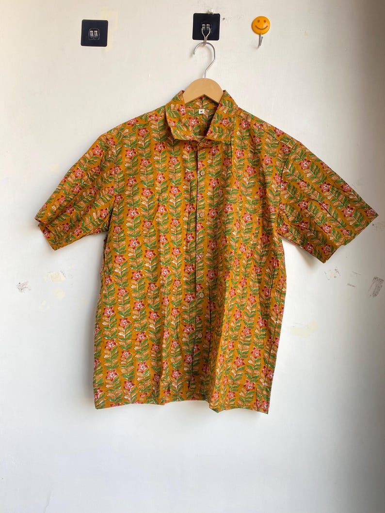 May include: A short-sleeved button-down shirt in a mustard yellow color, adorned with a repeating floral pattern in pink and green. The shirt is hanging on a wooden hanger against a white wall.