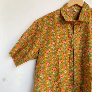 May include: Mustard yellow short-sleeved shirt with a floral pattern. The shirt features a repeating design of pink flowers and green leaves on a mustard yellow background. It has a collared neckline and wooden buttons. The shirt is hanging on a wooden hanger.
