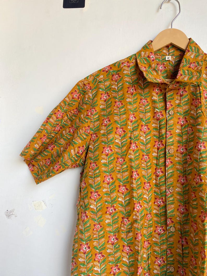 May include: Mustard yellow short-sleeved shirt with a floral pattern. The shirt features a repeating design of pink flowers and green leaves on a mustard yellow background. It has a collared neckline and wooden buttons. The shirt is hanging on a wooden hanger.