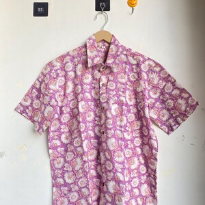 May include: Short-sleeved button-down shirt with a floral pattern in shades of purple, pink, and white. The shirt is hanging on a wooden hanger against a white wall. The floral design covers the entire shirt.