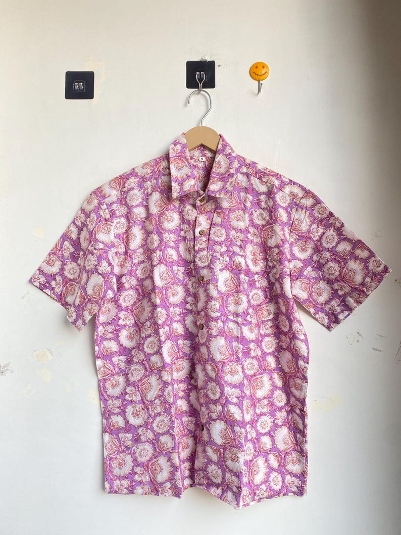 May include: Short-sleeved button-down shirt with a floral pattern in shades of purple, pink, and white. The shirt is hanging on a wooden hanger against a white wall. The floral design covers the entire shirt.