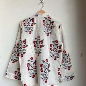 May include: Cream-colored long-sleeved shirt with a floral pattern. The design features red flowers and blue leaves on a white background. The shirt is hanging on a wooden hanger.