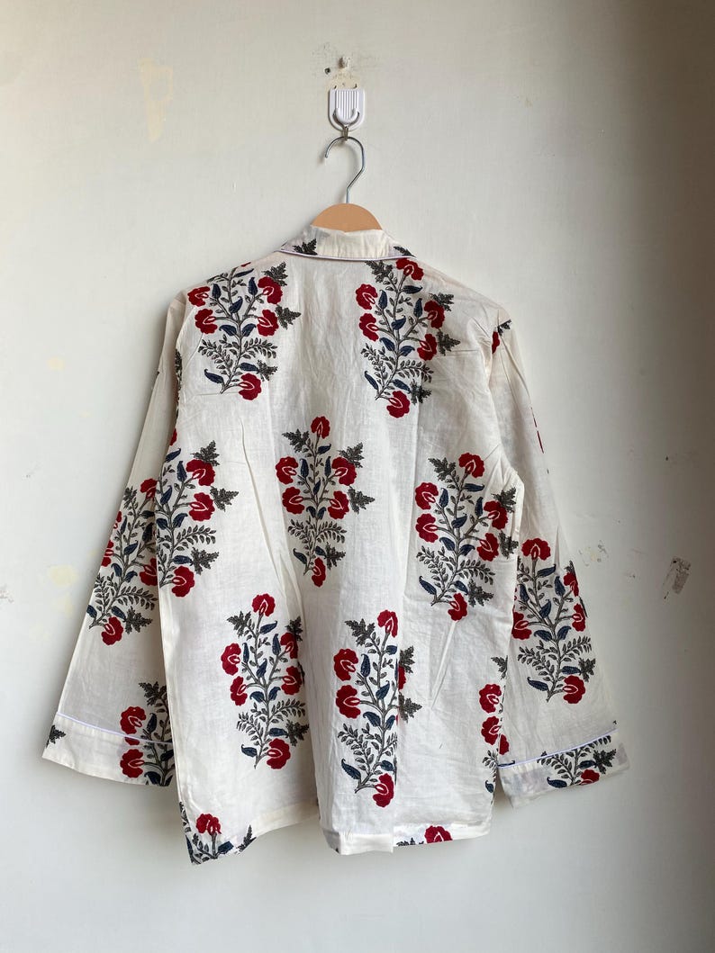 May include: Cream-colored long-sleeved shirt with a floral pattern. The design features red flowers and blue leaves on a white background. The shirt is hanging on a wooden hanger.