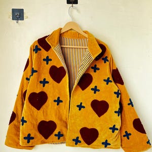 May include: Mustard yellow jacket with a velvet texture, decorated with large burgundy hearts and blue cross accents. The jacket has a collared neckline and a striped interior lining. It is hanging on a wooden hanger.