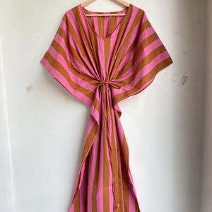 May include: A pink and brown striped kaftan dress hanging on a wooden hanger. The dress has a V-neck and a tie at the waist. The fabric is a lightweight material, and the dress has a loose, flowing silhouette.