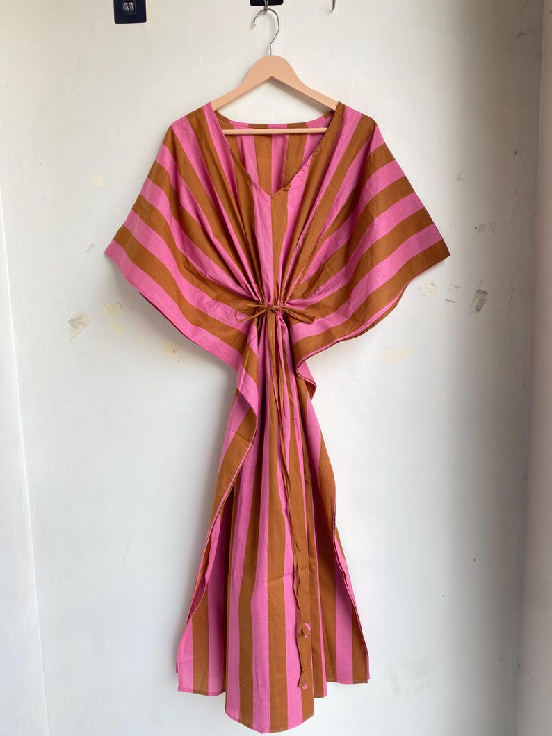 May include: A pink and brown striped kaftan dress hanging on a wooden hanger. The dress has a V-neck and a tie at the waist. The fabric is a lightweight material, and the dress has a loose, flowing silhouette.