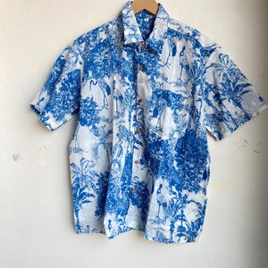 May include: Short-sleeved button-down shirt with a blue and white tropical print featuring birds and foliage. The shirt has a classic collar and wooden buttons. The fabric appears lightweight and suitable for warm weather.