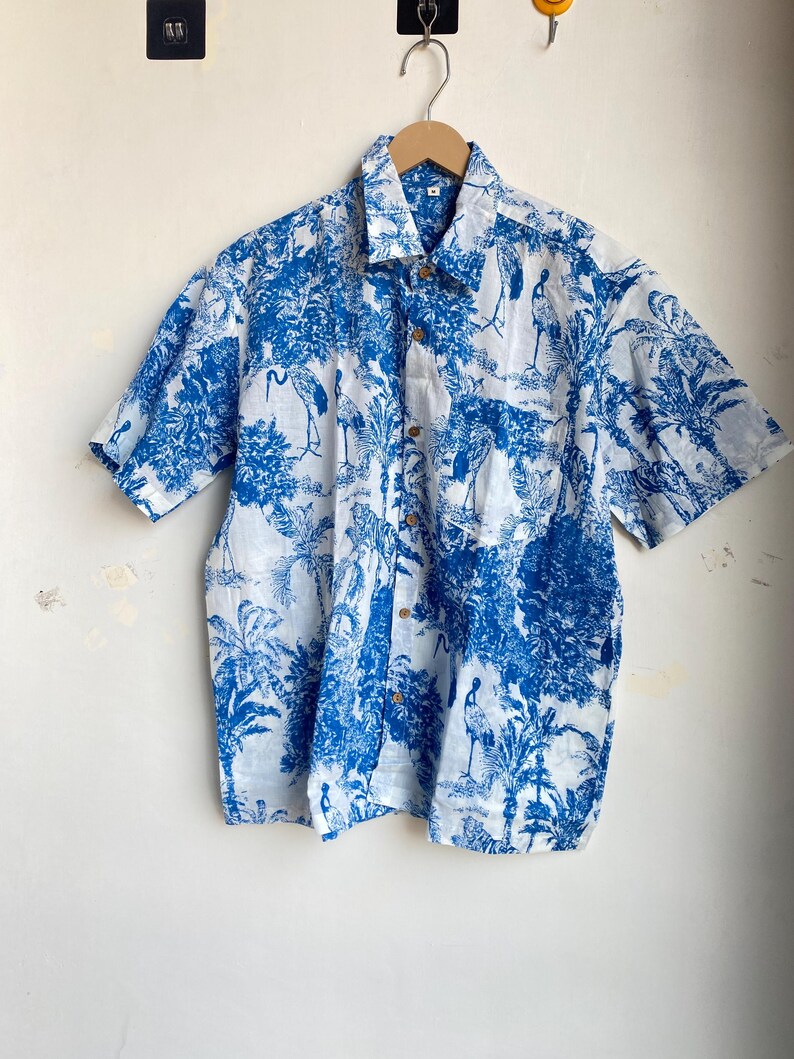 May include: Short-sleeved button-down shirt with a blue and white tropical print featuring birds and foliage. The shirt has a classic collar and wooden buttons. The fabric appears lightweight and suitable for warm weather.