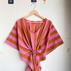 May include: A pink and brown striped kaftan dress with a V-neck and tie waist. The dress is hanging on a wooden hanger against a white wall. The kaftan has wide sleeves and a loose, flowing design.