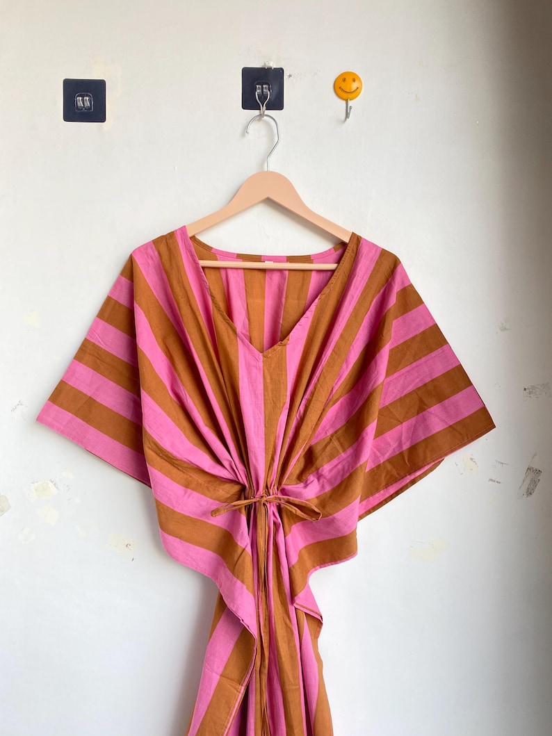 May include: A pink and brown striped kaftan dress with a V-neck and tie waist. The dress is hanging on a wooden hanger against a white wall. The kaftan has wide sleeves and a loose, flowing design.