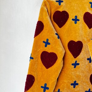 May include: Mustard yellow jacket with a velvet texture, decorated with large burgundy hearts and blue cross patterns. The jacket has a collared neckline and is hanging on a wooden hanger.