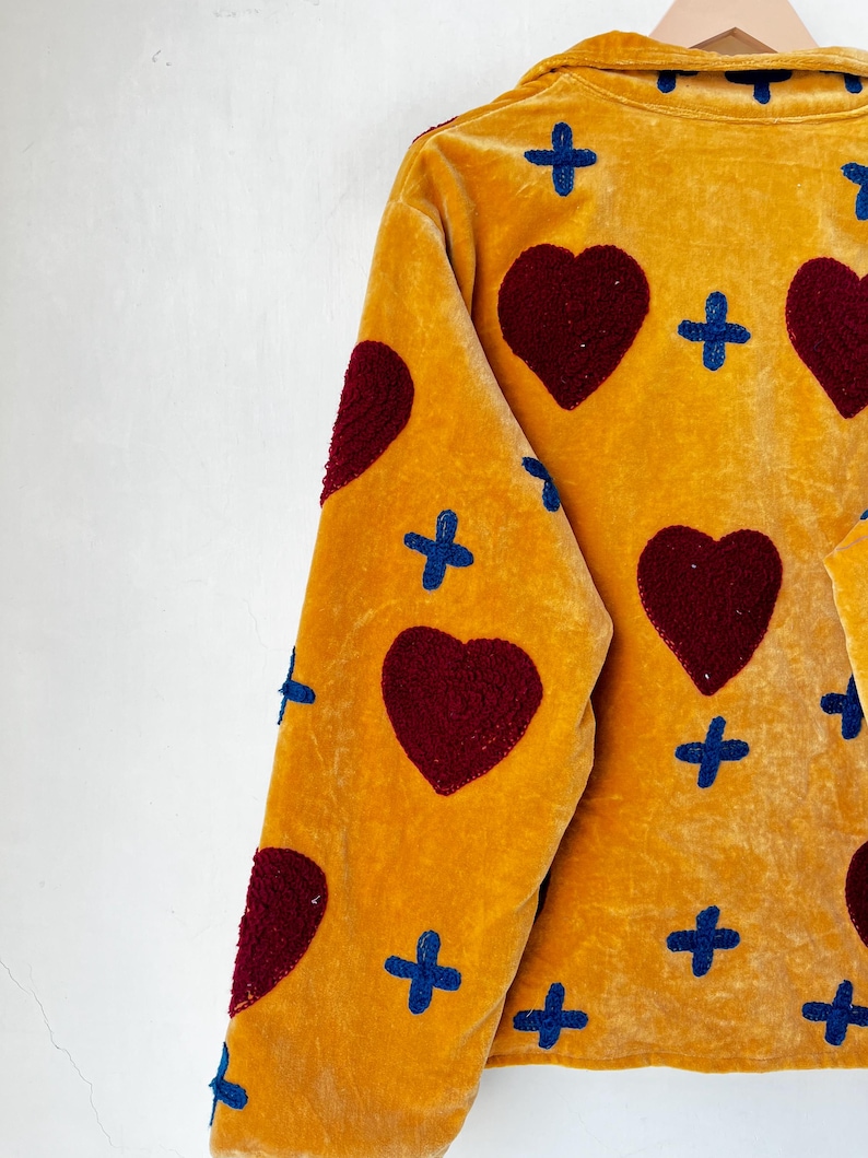 May include: Mustard yellow jacket with a velvet texture, decorated with large burgundy hearts and blue cross patterns. The jacket has a collared neckline and is hanging on a wooden hanger.