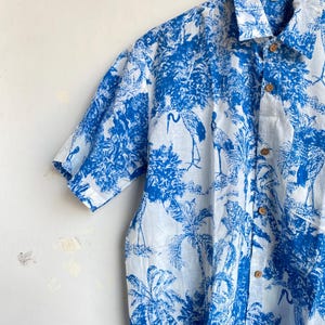 May include: A short-sleeved button-down shirt with a blue and white tropical print featuring cranes, trees, and other foliage. The shirt has wooden buttons and a classic collar. The fabric appears to be lightweight.