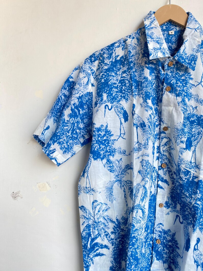 May include: A short-sleeved button-down shirt with a blue and white tropical print featuring cranes, trees, and other foliage. The shirt has wooden buttons and a classic collar. The fabric appears to be lightweight.