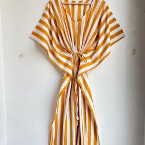May include: A mustard yellow and white striped kaftan dress hanging on a wooden hanger. The dress has a V-neckline and a drawstring waist. The fabric is lightweight and appears to be cotton or a similar material. The dress has wide sleeves and a flowing skirt.