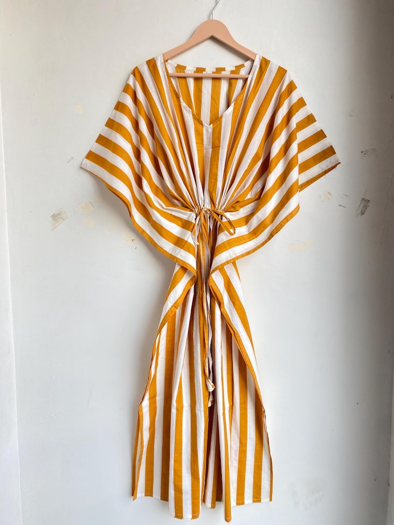 May include: A mustard yellow and white striped kaftan dress hanging on a wooden hanger. The dress has a V-neckline and a drawstring waist. The fabric is lightweight and appears to be cotton or a similar material. The dress has wide sleeves and a flowing skirt.