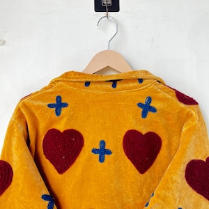 May include: Mustard yellow velvet jacket with embroidered burgundy hearts and blue crosses. The jacket is hanging on a wooden hanger against a white wall. The design is visible on the back of the jacket.
