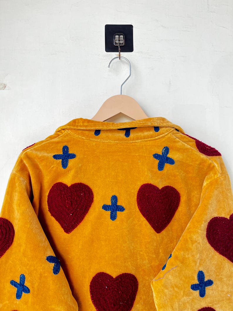 May include: Mustard yellow velvet jacket with embroidered burgundy hearts and blue crosses. The jacket is hanging on a wooden hanger against a white wall. The design is visible on the back of the jacket.