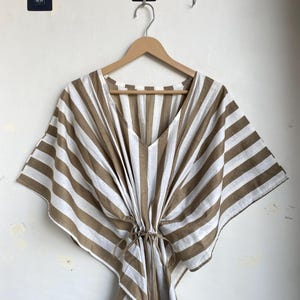 May include: A brown and white striped caftan dress with a V-neck and a drawstring waist. The dress is hanging on a wooden hanger against a white wall. The caftan has wide sleeves and a loose fit.
