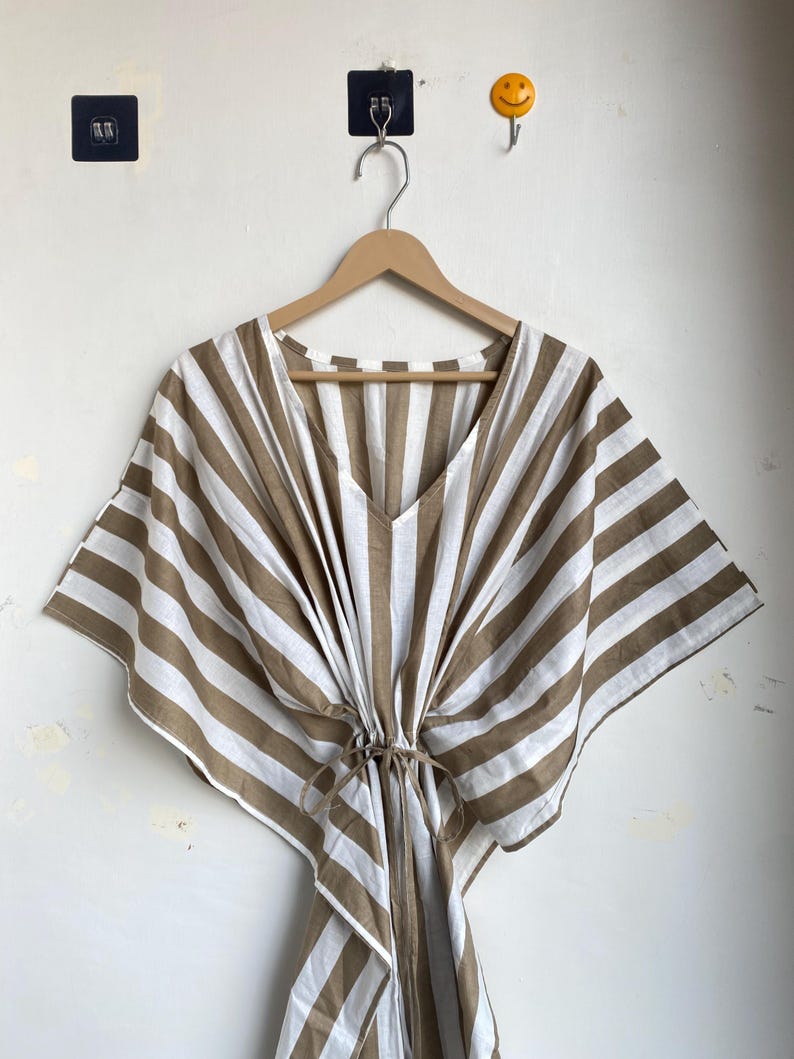 May include: A brown and white striped caftan dress with a V-neck and a drawstring waist. The dress is hanging on a wooden hanger against a white wall. The caftan has wide sleeves and a loose fit.