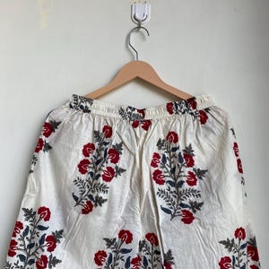 May include: White palazzo pants with a red floral pattern and blue accents. The pants are hanging on a wooden hanger. The floral design is repeated throughout the fabric.