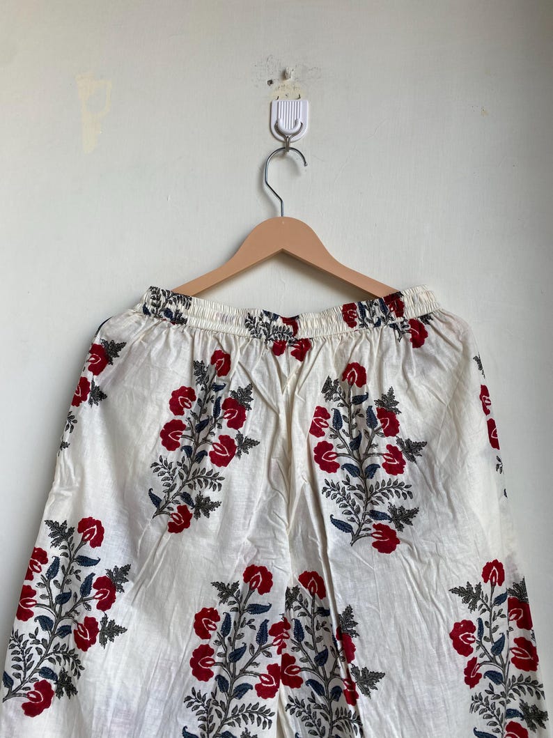 May include: White palazzo pants with a red floral pattern and blue accents. The pants are hanging on a wooden hanger. The floral design is repeated throughout the fabric.