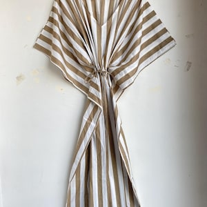 May include: A striped, brown and white caftan dress with a V-neck and tie waist, hanging on a wooden hanger. The dress features wide, vertical stripes and a loose, flowing design, perfect for warm weather.