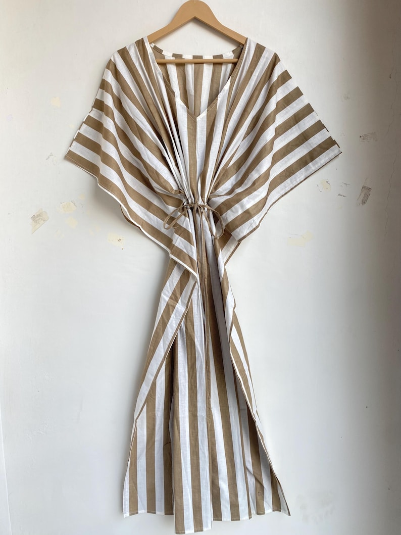 May include: A striped, brown and white caftan dress with a V-neck and tie waist, hanging on a wooden hanger. The dress features wide, vertical stripes and a loose, flowing design, perfect for warm weather.