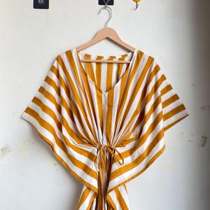 May include: A yellow and white striped kaftan dress with a V-neck and tie waist. The dress is hanging on a wooden hanger against a white wall. The kaftan has wide sleeves and a loose, flowing fit.