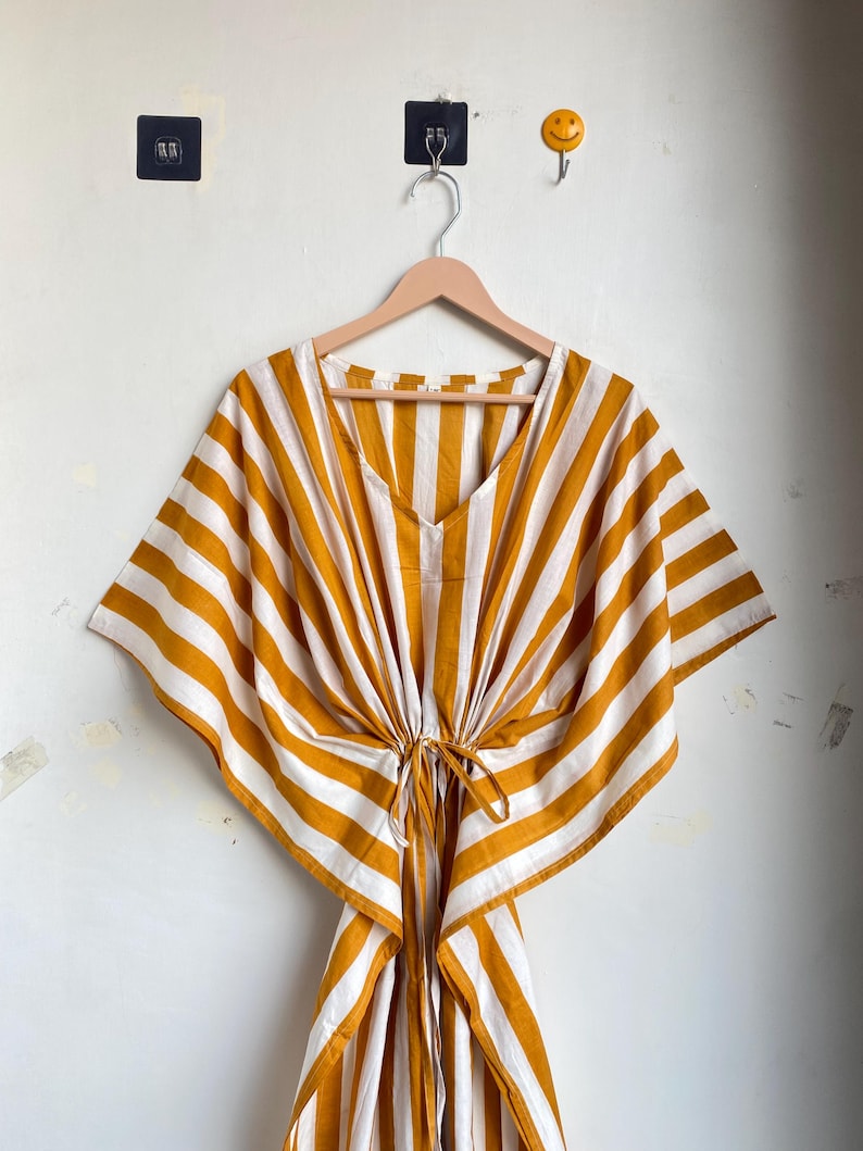 May include: A yellow and white striped kaftan dress with a V-neck and tie waist. The dress is hanging on a wooden hanger against a white wall. The kaftan has wide sleeves and a loose, flowing fit.