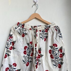 May include: White palazzo pants with a red floral pattern and blue-green leaves. The pants are hanging on a wooden hanger against a white wall. The floral design is repeated throughout the fabric.