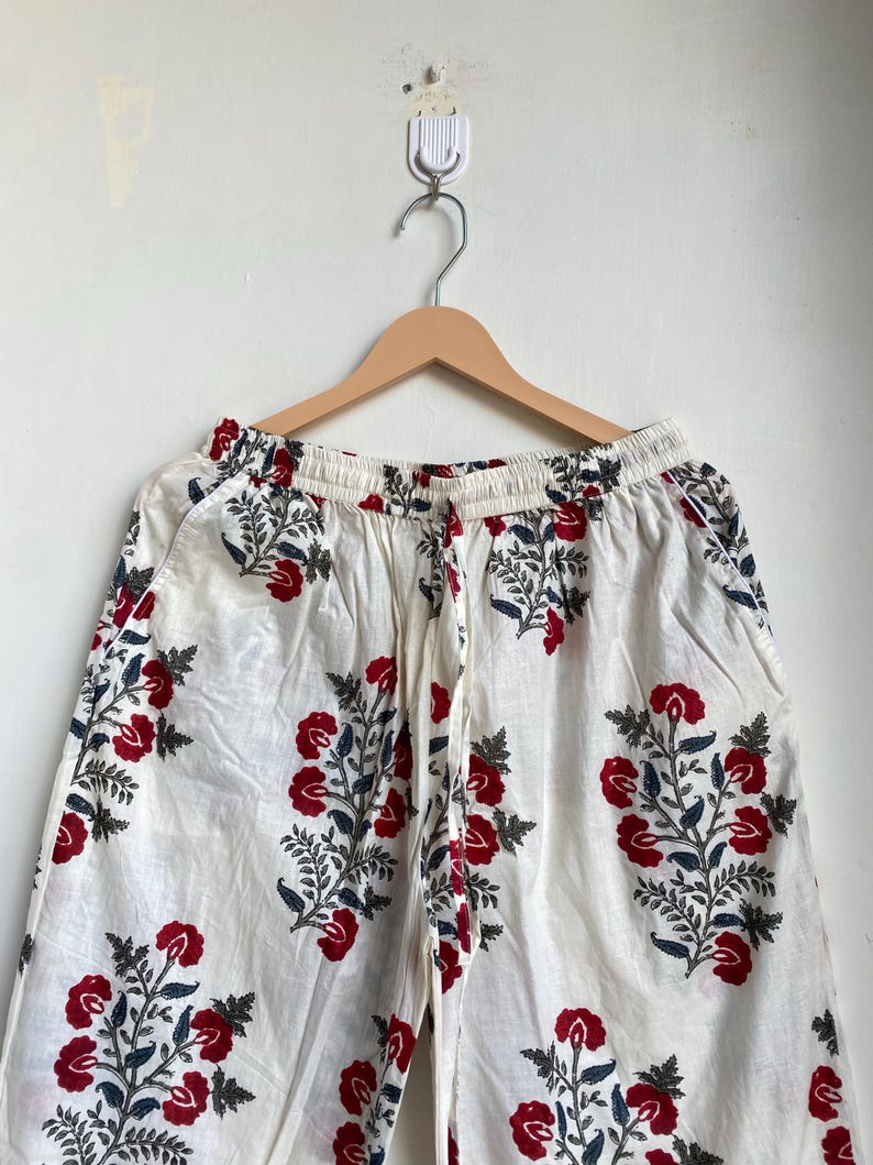 May include: White palazzo pants with a red floral pattern and blue-green leaves. The pants are hanging on a wooden hanger against a white wall. The floral design is repeated throughout the fabric.