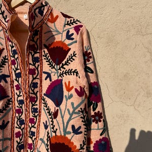 May include: A peach-colored jacket with a floral embroidery design. The embroidery features a variety of colors, including red, orange, blue, green, and purple. The jacket has a stand-up collar and a zipper closure.