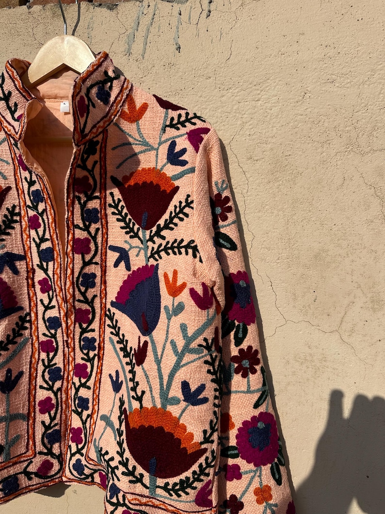 May include: A peach-colored jacket with a floral embroidery design. The embroidery features a variety of colors, including red, orange, blue, green, and purple. The jacket has a stand-up collar and a zipper closure.