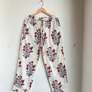 May include: White pajama pants with a red and blue floral pattern. The pants are hanging on a wooden hanger against a white wall. The floral design is repeated across the fabric.