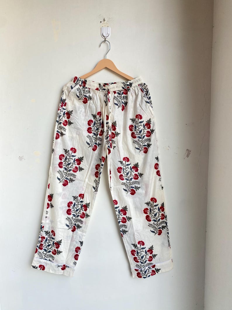 May include: White pajama pants with a red and blue floral pattern. The pants are hanging on a wooden hanger against a white wall. The floral design is repeated across the fabric.