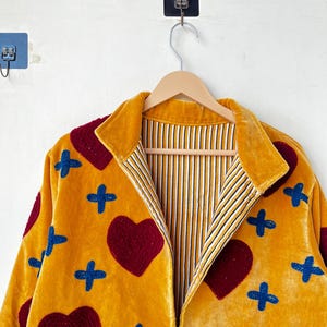 May include: Mustard yellow velvet jacket with a collared neckline. The jacket features appliqued burgundy hearts and blue cross shapes. The interior lining has a striped pattern of white, yellow, and black. The jacket is hanging on a wooden hanger.