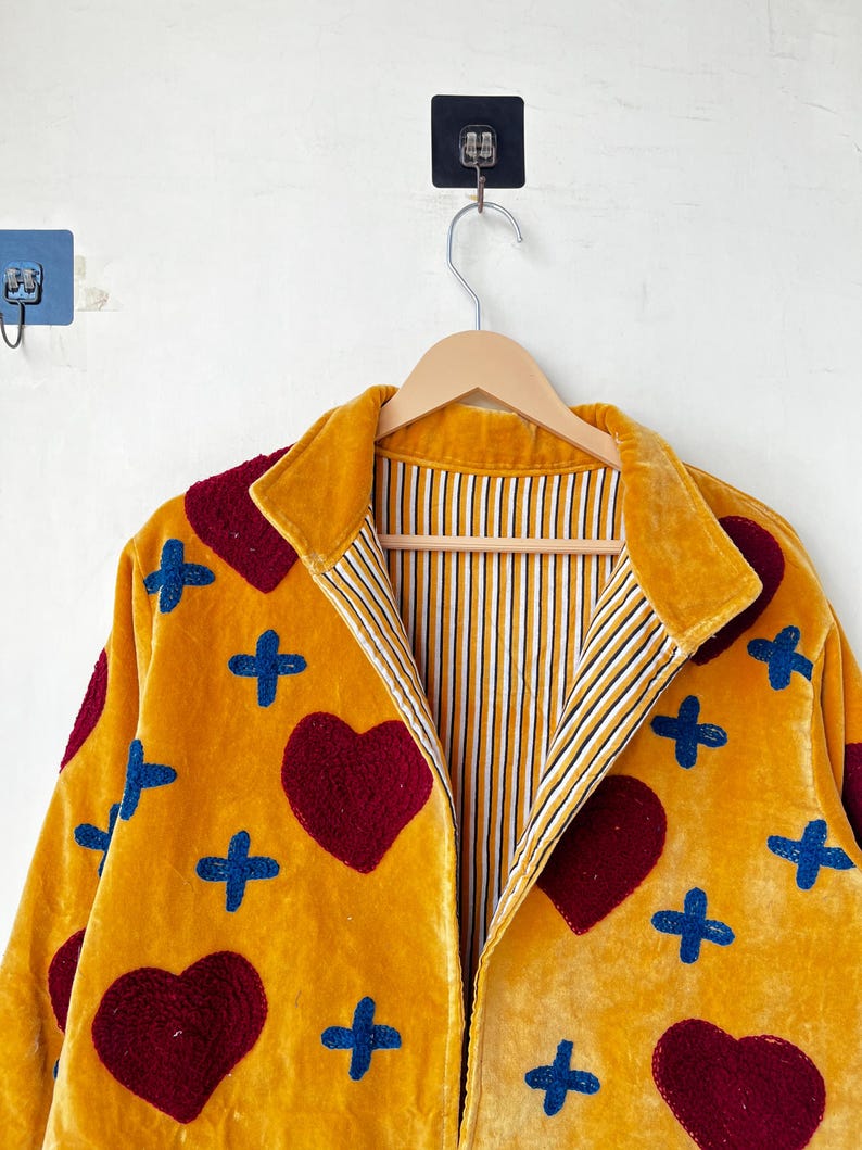 May include: Mustard yellow velvet jacket with a collared neckline. The jacket features appliqued burgundy hearts and blue cross shapes. The interior lining has a striped pattern of white, yellow, and black. The jacket is hanging on a wooden hanger.