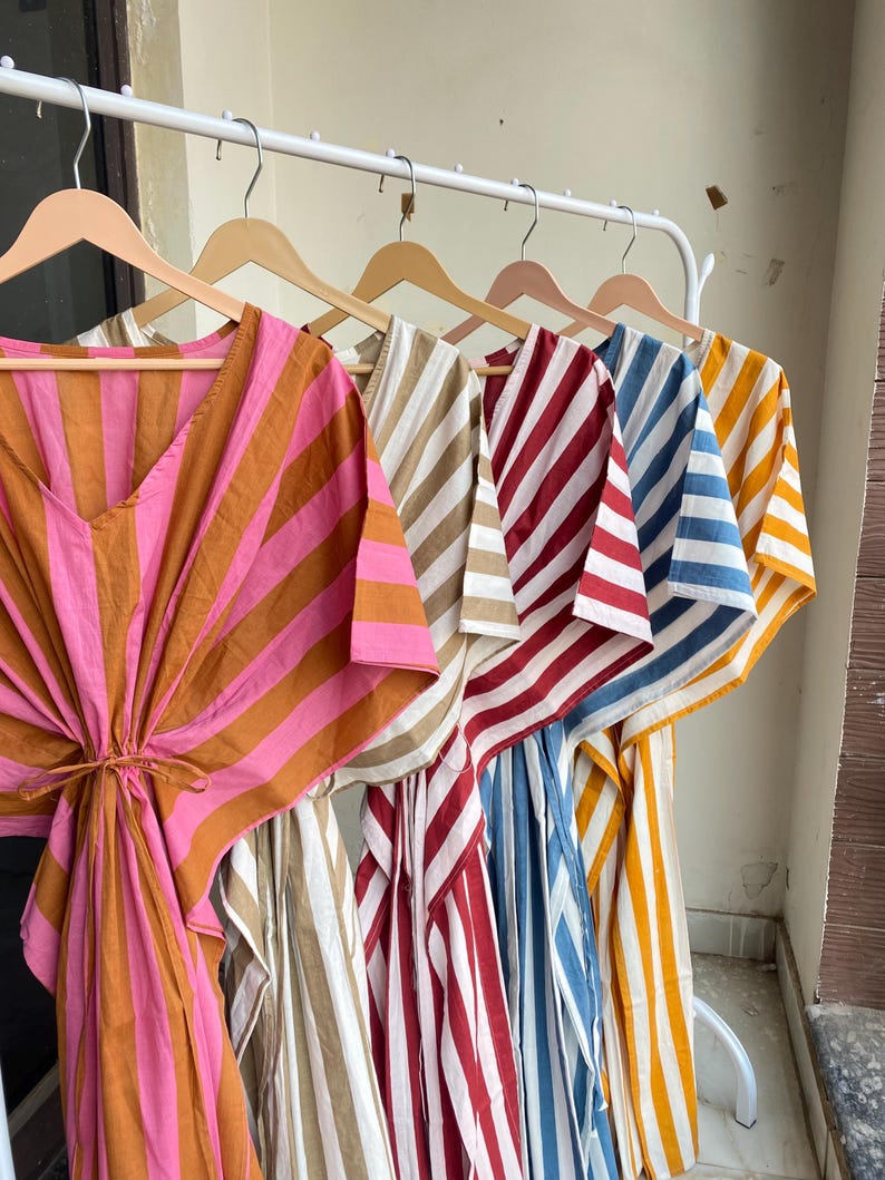May include: A row of colorful, striped caftans hanging on wooden hangers. The caftans feature bold stripes in various colors, including pink and orange, beige and white, red and white, blue and white, and yellow and white. The caftans have a loose, flowing design.