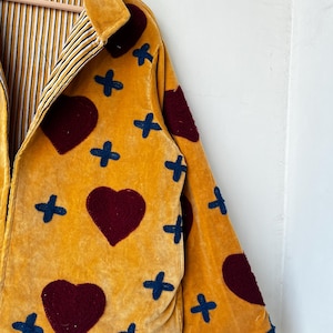 May include: Mustard yellow jacket with burgundy heart and navy blue cross appliqu&eacute;s. The jacket has a collared neckline and a striped lining in yellow, black, and white. The jacket is made of a soft, velvety material.