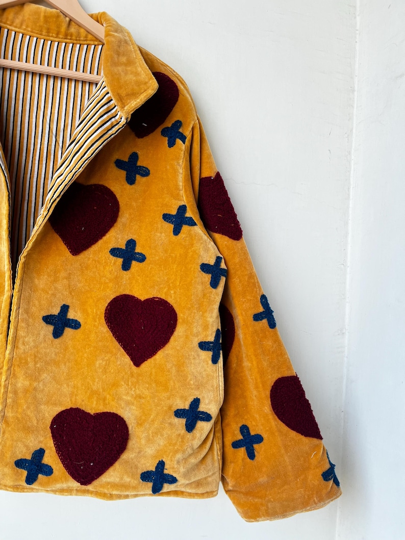May include: Mustard yellow jacket with burgundy heart and navy blue cross appliqu&eacute;s. The jacket has a collared neckline and a striped lining in yellow, black, and white. The jacket is made of a soft, velvety material.