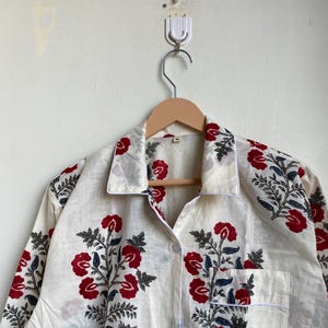 May include: A white button-down shirt with a floral pattern of red flowers and blue leaves. The shirt has a pocket on the front and is hanging on a wooden hanger. The shirt is made of a lightweight fabric.
