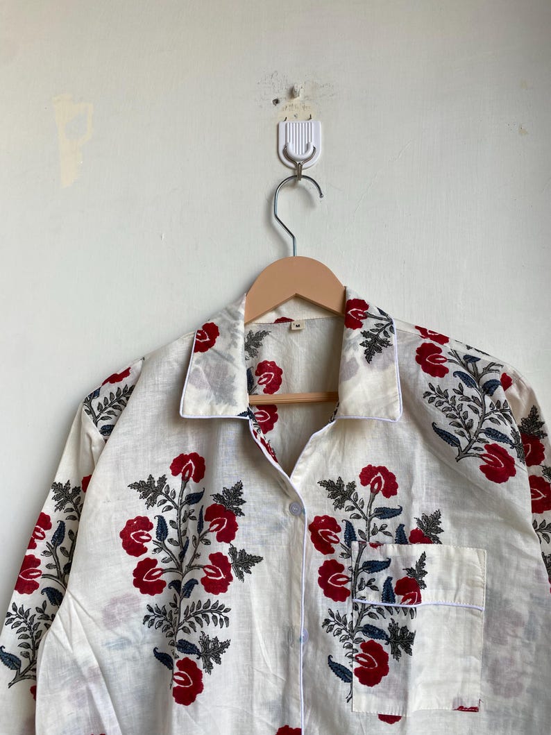 May include: A white button-down shirt with a floral pattern of red flowers and blue leaves. The shirt has a pocket on the front and is hanging on a wooden hanger. The shirt is made of a lightweight fabric.