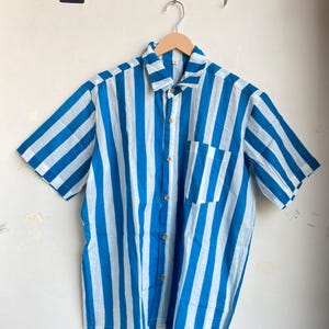 May include: Short-sleeved button-down shirt with vertical blue and white stripes. The shirt has a single chest pocket and is hanging on a wooden hanger. The shirt is made of a lightweight fabric.