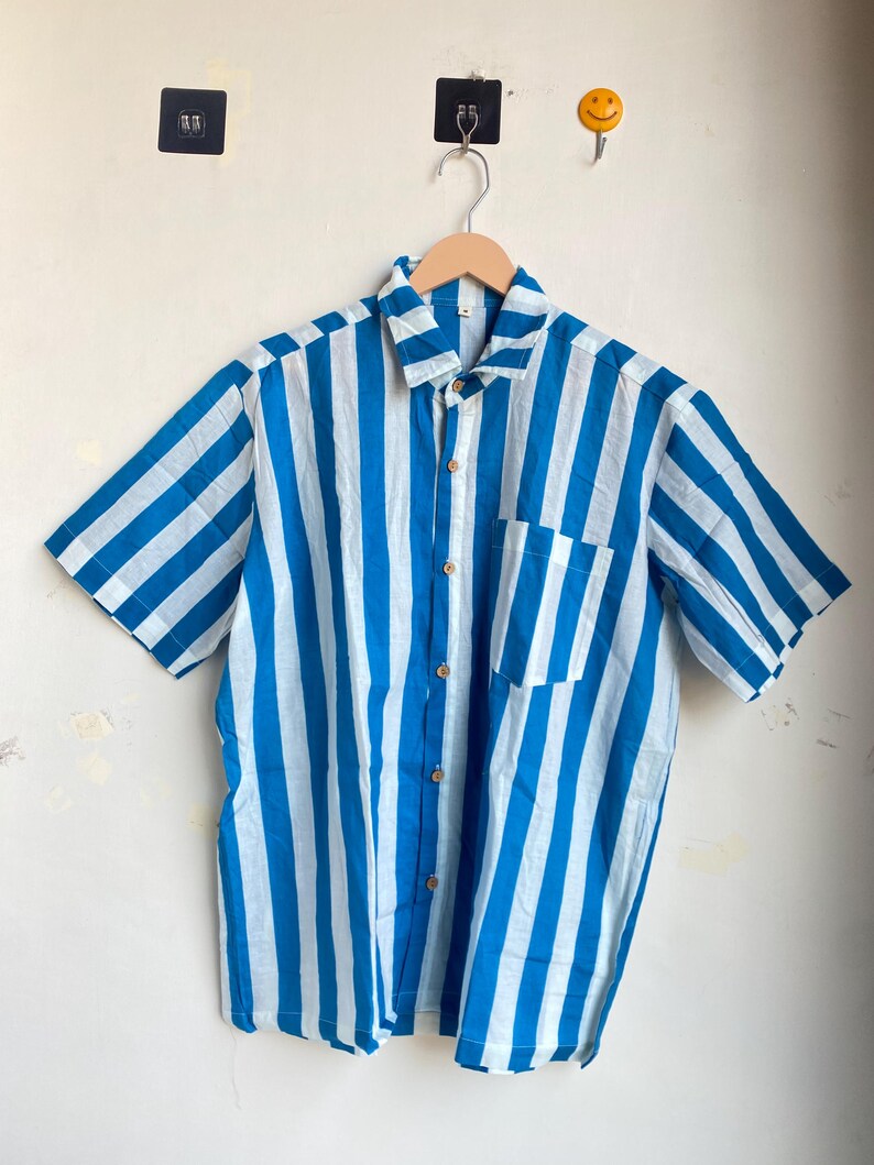 May include: Short-sleeved button-down shirt with vertical blue and white stripes. The shirt has a single chest pocket and is hanging on a wooden hanger. The shirt is made of a lightweight fabric.