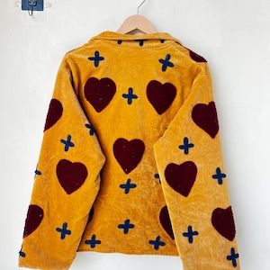 May include: Mustard yellow jacket with a velvet texture, featuring large burgundy heart appliqu&eacute;s and navy blue cross accents. The jacket is hanging on a wooden hanger against a white wall.